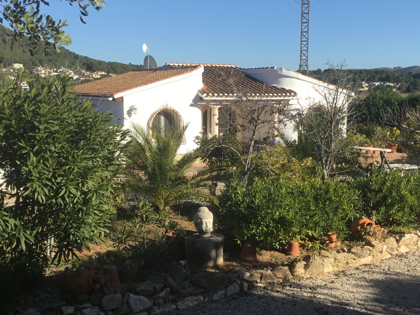 Vue extérieure de la villa méditerranéenne aux murs blancs, fenêtres en arc, toit en tuiles et jardin paysager avec palmiers et statue de Bouddha.