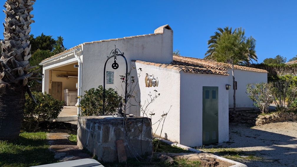 Vue extérieure d'une dépendance méditerranéenne traditionnelle avec façade blanche, toit en tuiles et un puits décoratif en pierre orné de fer forgé.