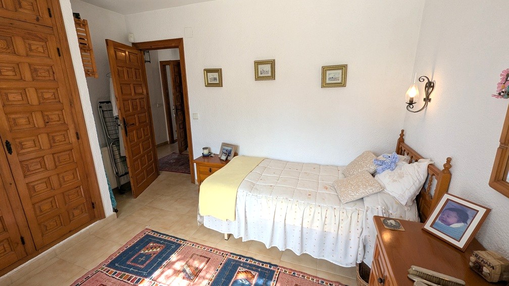 Chambre à coucher avec placards intégrés en bois sculpté, sol en carrelage et lit simple. Elle dispose de menuiseries traditionnelles et de murs blancs texturés.