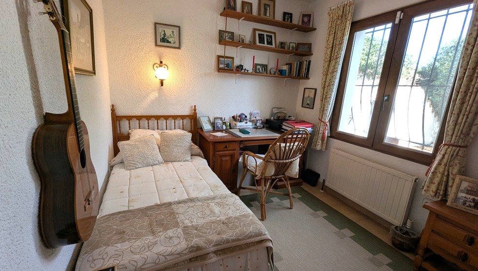 Chambre avec lit simple en bois, bureau classique, chaise en osier et étagères murales. Comprend un radiateur et une fenêtre sécurisée par des grilles.