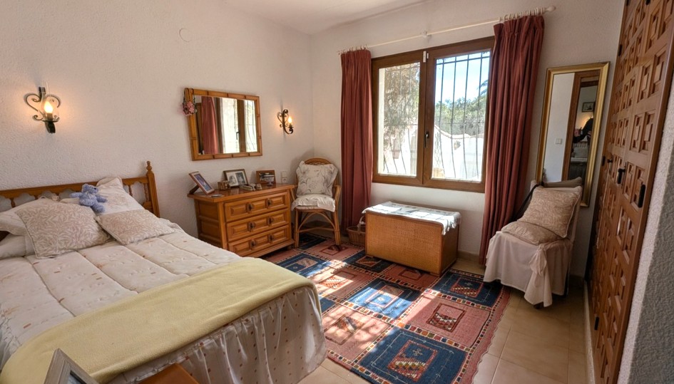 Chambre de style traditionnel avec placards intégrés en bois, commode assortie et lumière naturelle. Murs texturés et sol en carrelage avec tapis à motifs.