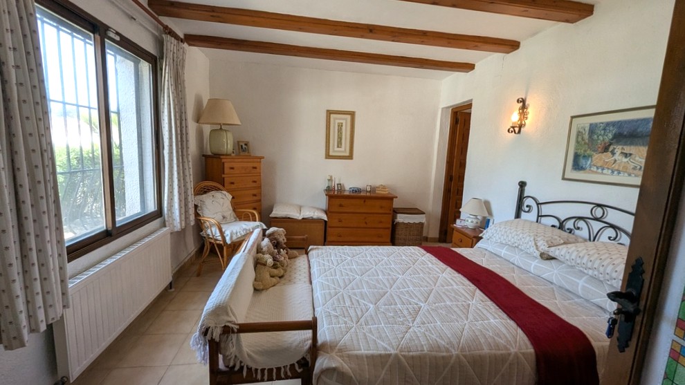 Chambre traditionnelle avec poutres en bois apparentes, cadre de lit en fer forgé et mobilier en bois. La pièce bénéficie d'une lumière naturelle et d'un radiateur.