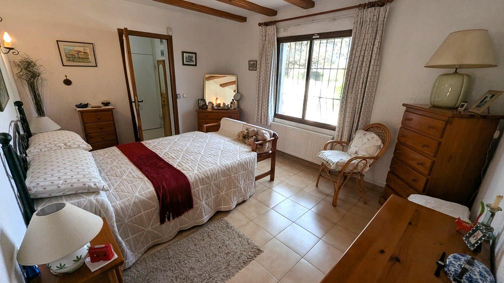 Chambre traditionnelle avec poutres en bois apparentes, sol en carrelage et radiateur sous la fenêtre apportant une luminosité naturelle constante.