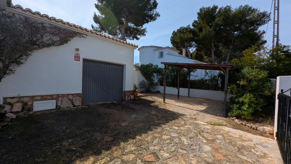 Garage avec porte métallique grise et abri de voiture en bois, entourés d'une allée en pierre et d'une façade méditerranéenne.