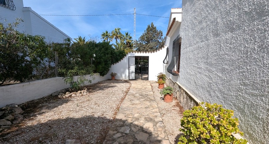Une allée pavée en pierre traverse une cour de gravier, bordée de murs blancs traditionnels, de plantes en pots et de grilles en fer forgé.