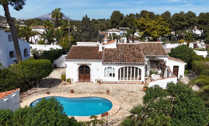 Vue aérienne de la villa avec piscine en forme de haricot, terrasse pavée et toiture en terre cuite, offrant une vue sur le mont Montgó.