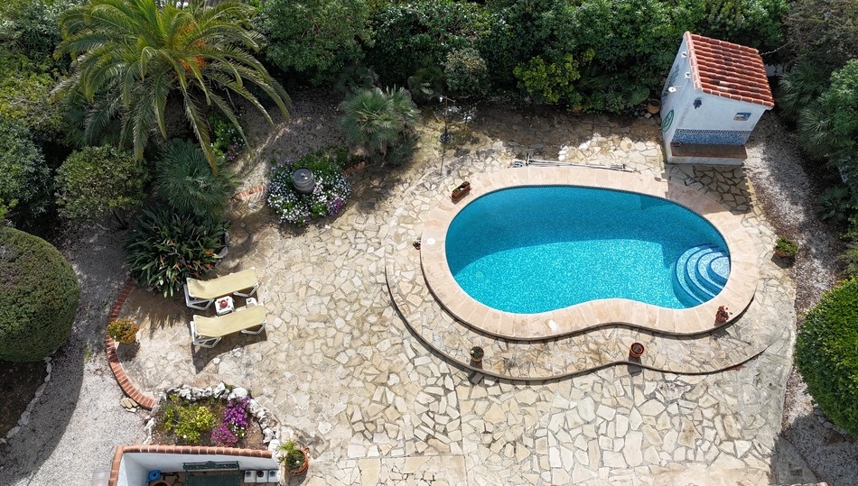 Vue aérienne d'une piscine de forme libre avec mosaïque bleue, entourée d'une terrasse en pierre naturelle et d'un jardin méditerranéen arboré de palmiers et d'arbustes.
