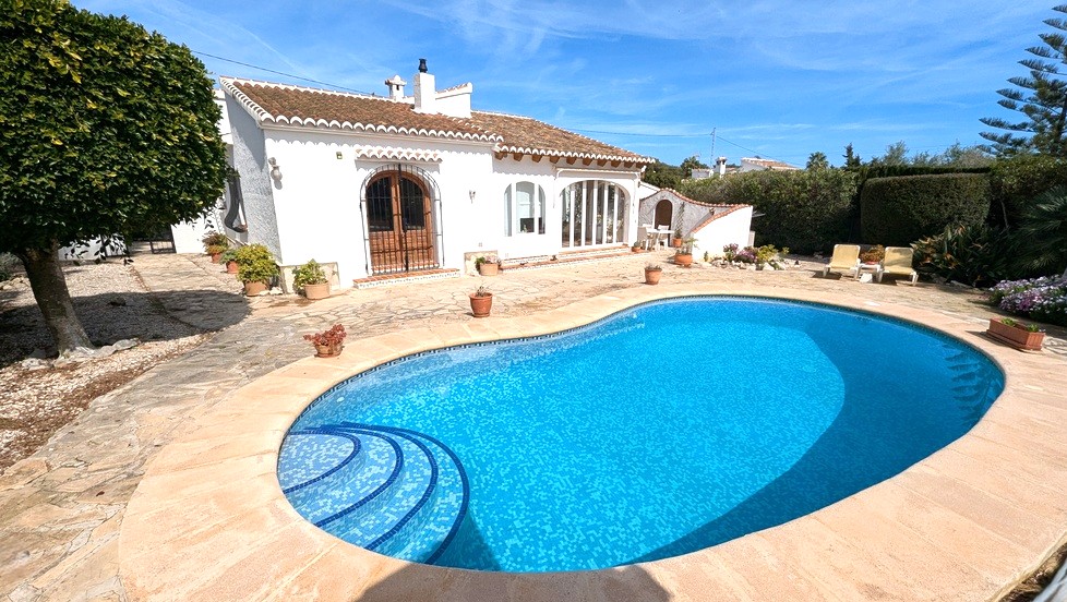 Vue extérieure de la piscine de forme libre avec mosaïque bleue, entourée d'une terrasse en pierre et de la villa méditerranéenne aux murs blancs.