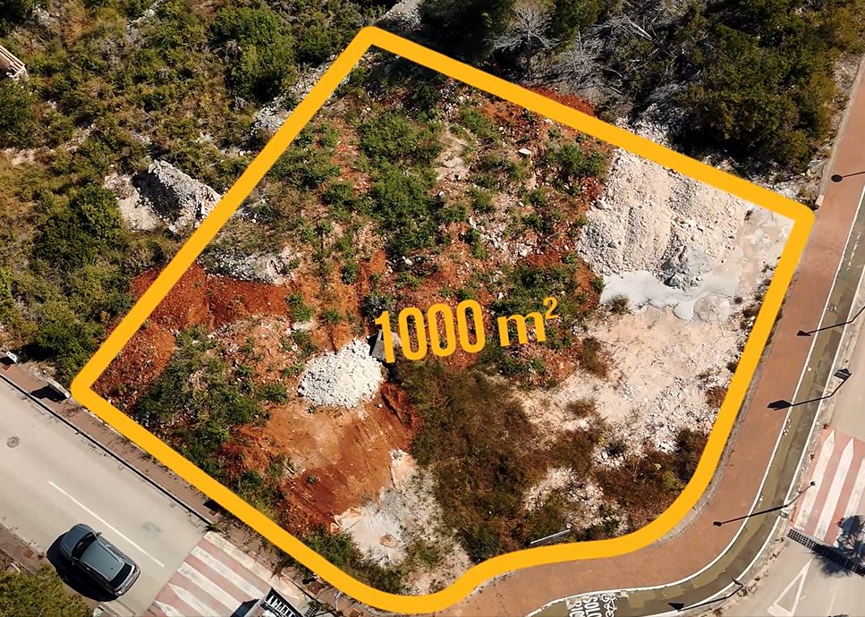 Vue aérienne de la parcelle d'angle de 1 000 m² à Jávea, montrant le terrain préparé avec son sol rouge caractéristique et les infrastructures routières adjacentes.