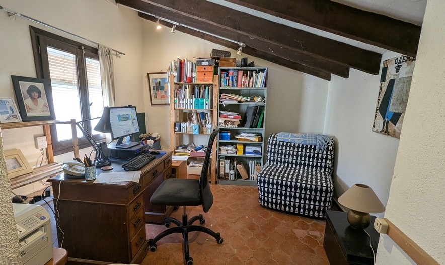 Bureau à domicile avec sol en terre cuite traditionnelle, poutres apparentes en bois sombre au plafond et bureau classique en bois sous une fenêtre latérale.