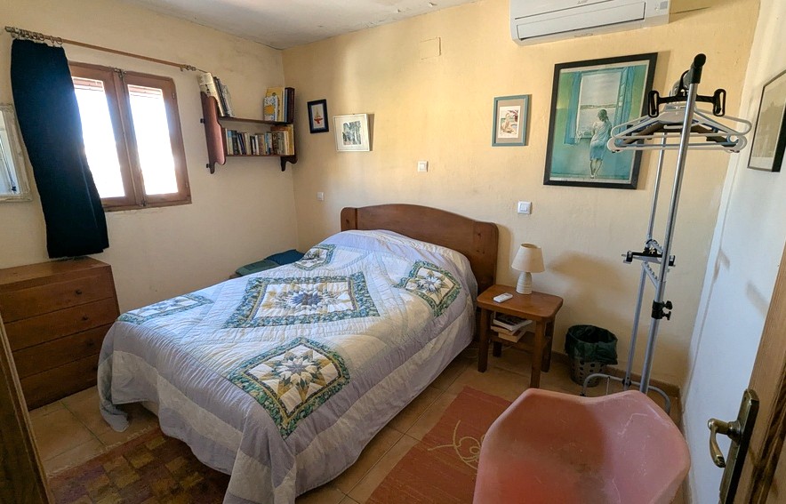 Chambre avec mobilier en bois, sol en carrelage et climatisation murale. Présente une étagère murale, une commode classique et une fenêtre en bois apportant de la lumière.