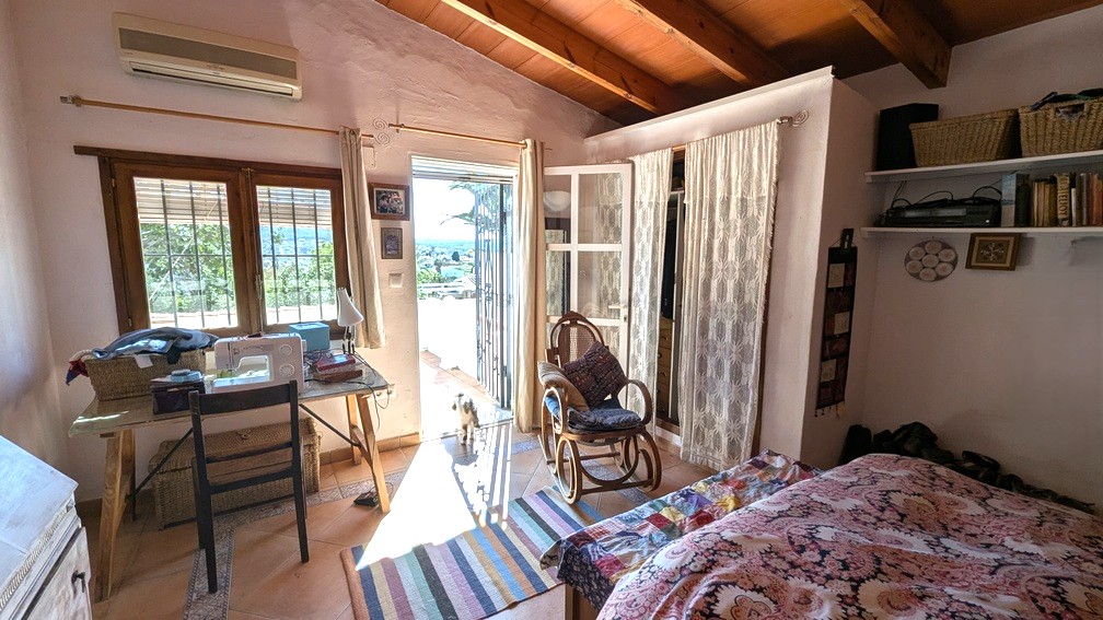 Chambre avec poutres en bois apparentes, sol en terre cuite et accès direct à une terrasse offrant une vue dégagée. Comprend la climatisation et un espace bureau.