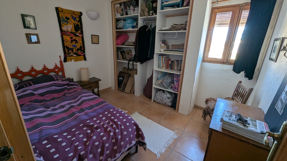 Chambre avec sol en terre cuite, étagères ouvertes intégrées et fenêtre en bois. L'espace comprend un lit simple et un bureau en bois sous une lumière naturelle.