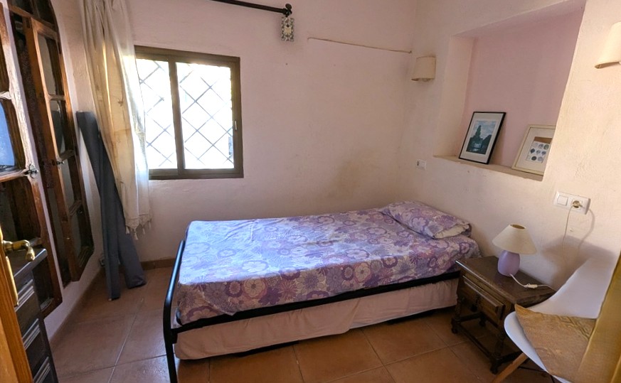 Chambre confortable avec lit simple, sol en terre cuite et niche murale décorative. Une fenêtre avec grilles de sécurité traditionnelles apporte de la lumière naturelle.