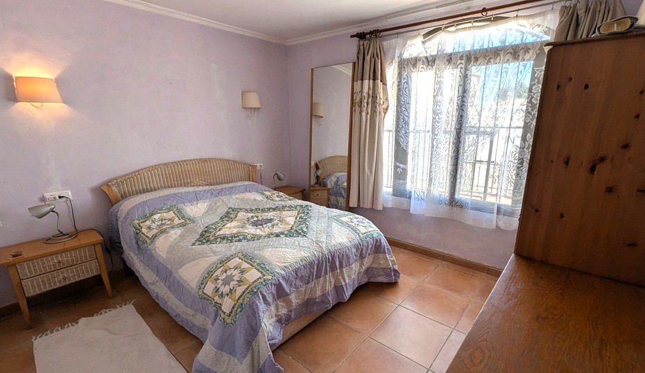 Chambre spacieuse aux murs lavande, sol en terre cuite traditionnelle, lit avec tête de lit en osier et grande fenêtre offrant une belle luminosité.