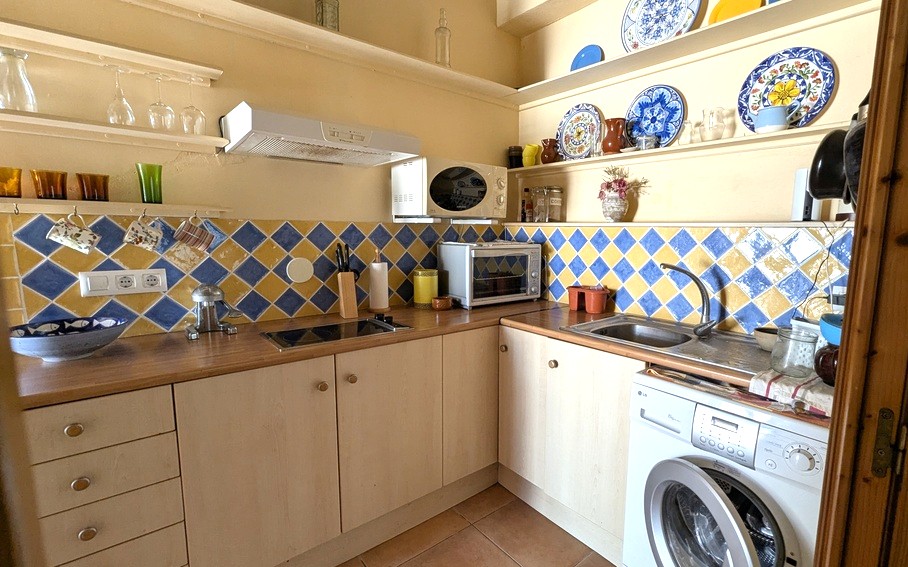 Cuisine compacte avec crédence en carreaux de losanges bleus et jaunes, plans de travail en bois et étagères ouvertes avec céramiques traditionnelles. Comprend un lave-linge.