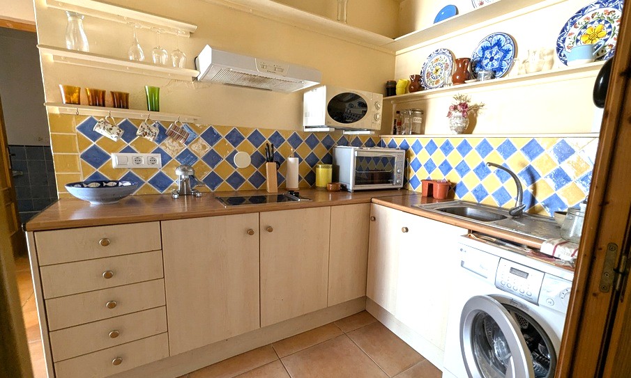 Cuisine compacte en L avec crédence en carreaux losanges bleus et jaunes, plans de travail en bois, étagères ouvertes et lave-linge intégré.
