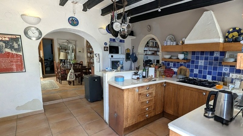 Cuisine traditionnelle avec meubles en bois, crédence en carreaux bleus et poutres apparentes sombres. Des arches mènent à la salle à manger attenante.