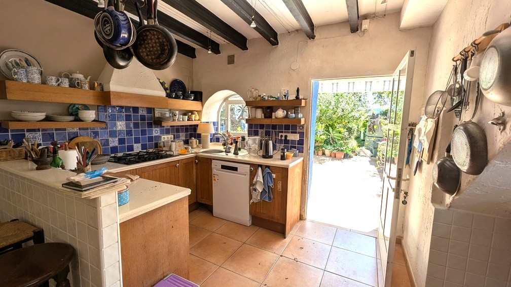 Cuisine traditionnelle avec poutres en bois sombre, crédence en carreaux bleus et accès direct à la terrasse ensoleillée bordée de végétation.