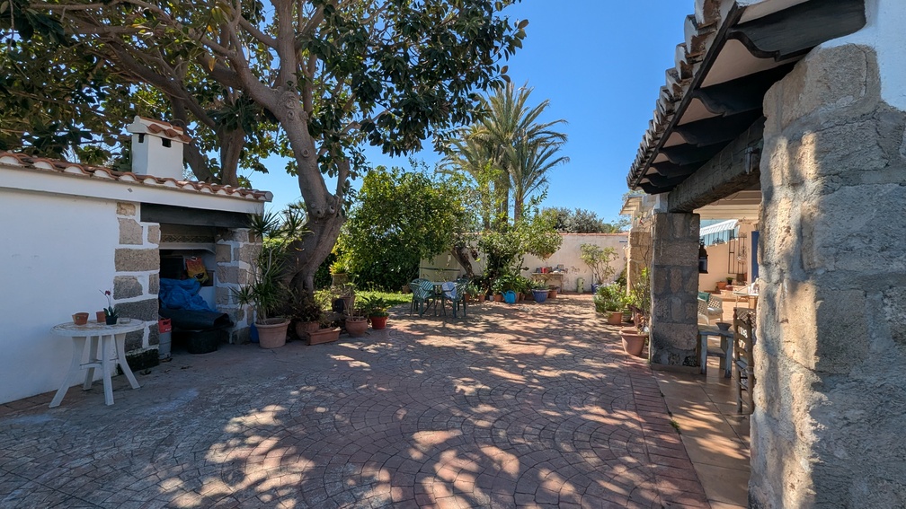Grand patio pavé avec piliers en pierre Tosca, naya ombragée et cuisine d'été extérieure, le tout entouré d'arbres matures et de plantes méditerranéennes.