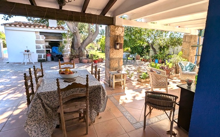 Naya orientée sud avec piliers en pierre Tosca, poutres en bois apparentes et coin repas extérieur donnant sur le jardin et la cuisine d'été.