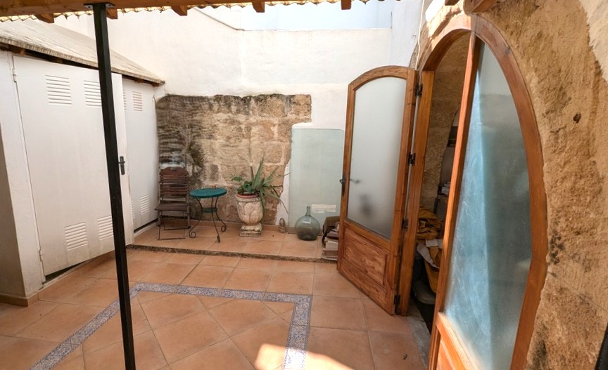 Patio traditionnel avec murs en pierre Tosca apparente, sol en terre cuite à bordures décoratives et portes cintrées en bois ouvrant sur l'intérieur.