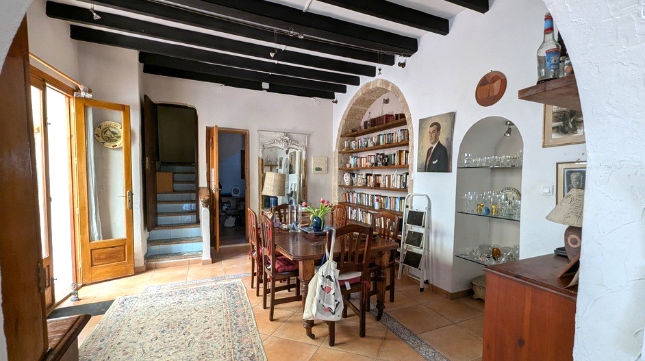 Salle à manger traditionnelle avec poutres apparentes sombres, bibliothèque intégrée sous une arche en pierre Tosca et sol en terre cuite menant à l'escalier.