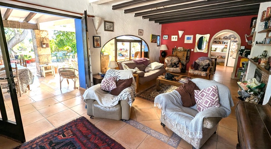 Salon spacieux doté de poutres en bois sombre, de sols en terre cuite et d'un mur rouge, s'ouvrant sur une naya traditionnelle et un jardin verdoyant.