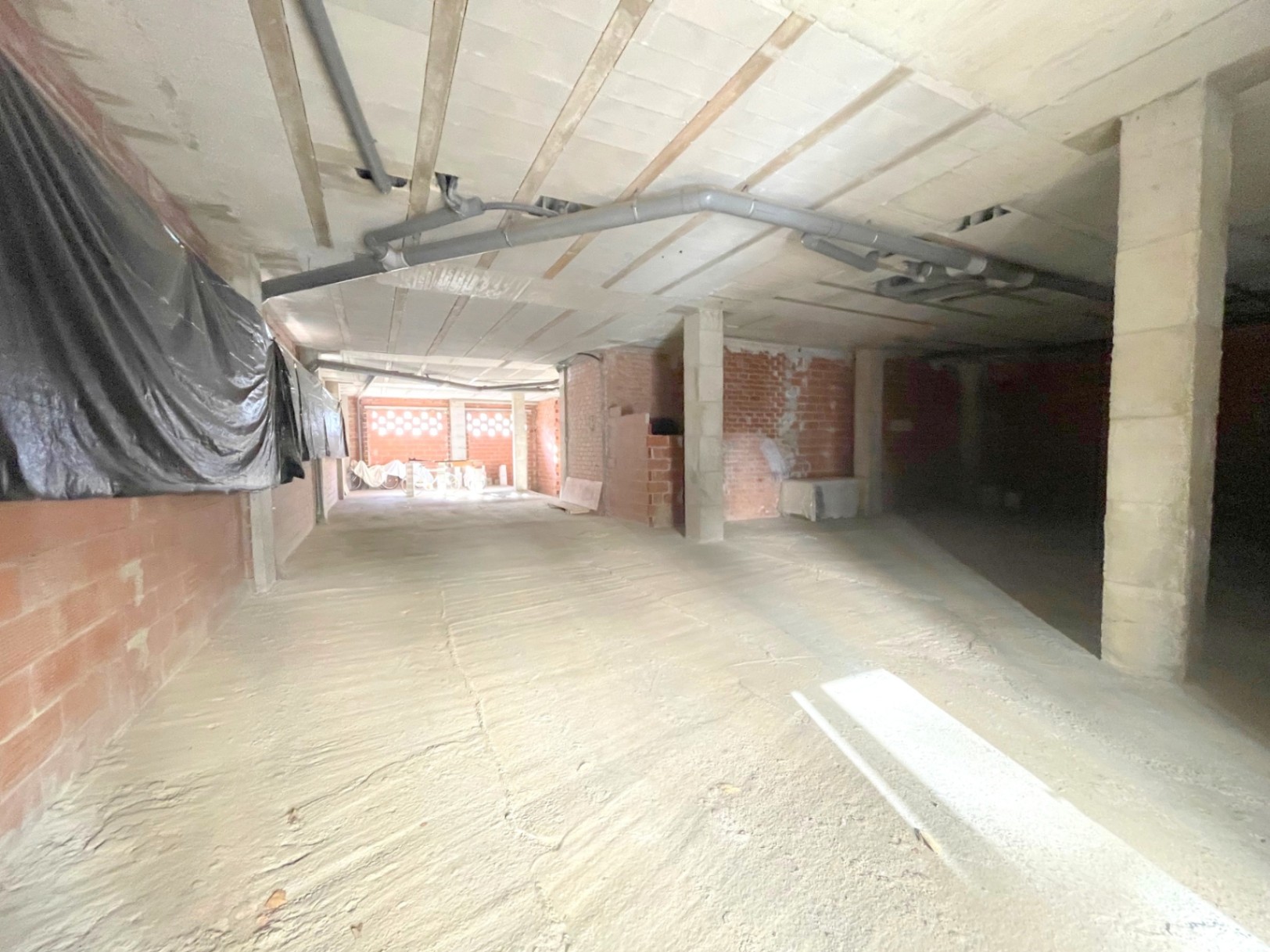 Espace commercial brut et ouvert avec piliers en béton, canalisations apparentes au plafond et murs en briques offrant un fort potentiel d'aménagement.