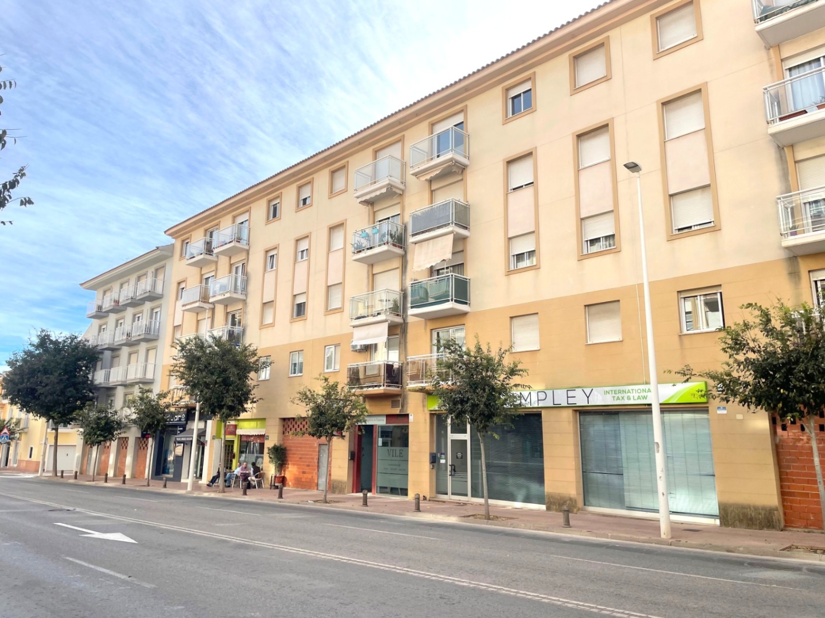 Façade extérieure d'un immeuble résidentiel avec locaux commerciaux au rez-de-chaussée, située sur une avenue arborée avec des balcons en verre.