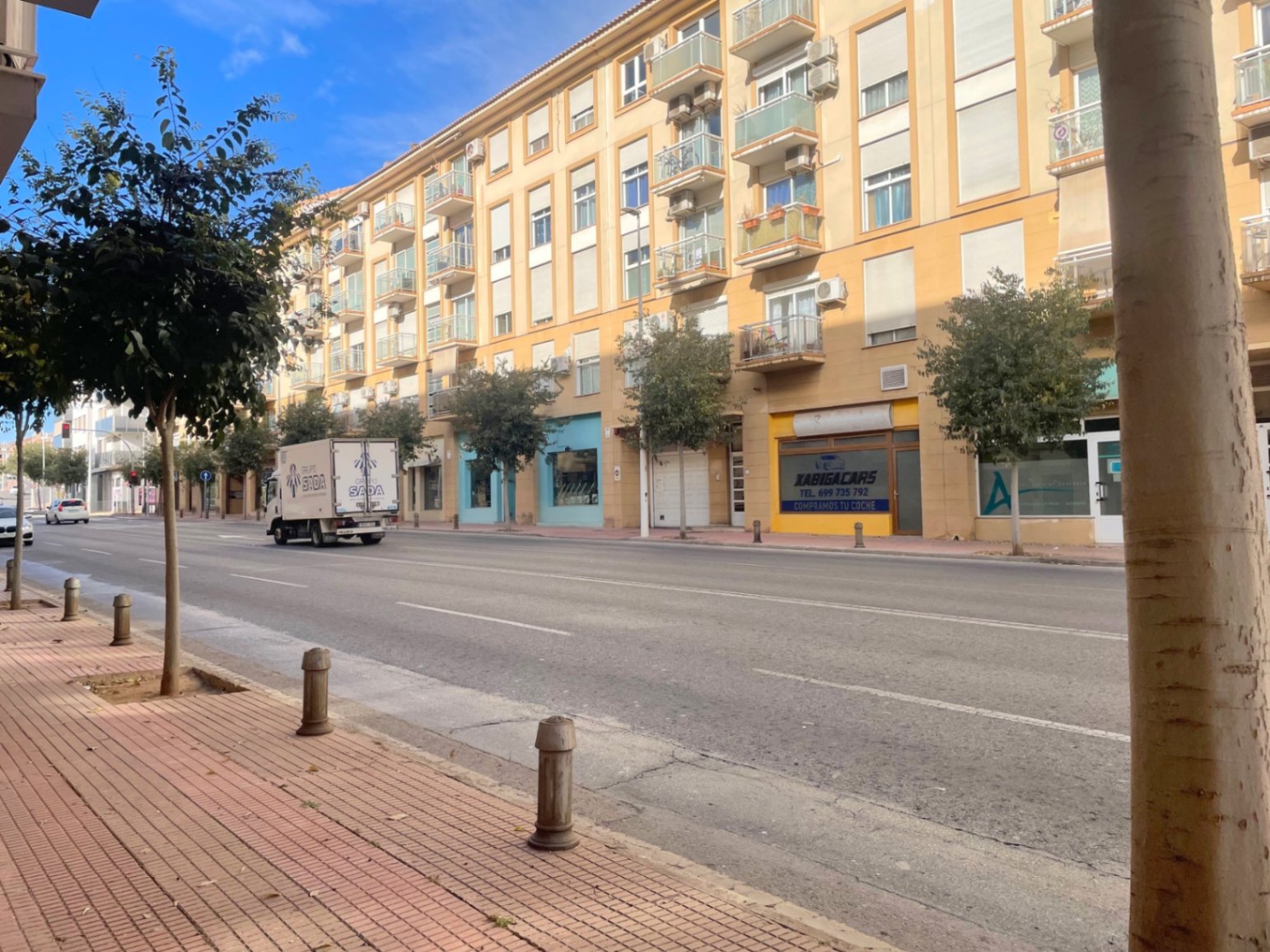 Vue extérieure de l'Avenida Palmela à Jávea, montrant la façade du bâtiment avec des locaux commerciaux au rez-de-chaussée et de larges trottoirs arborés.