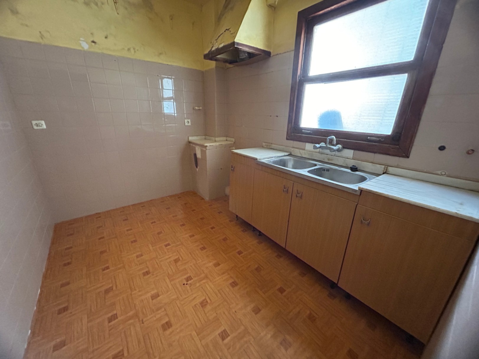 Cuisine d'origine avec évier double en inox, meubles en bois et carrelage à motifs. La fenêtre offre une lumière naturelle, idéale pour une rénovation complète.