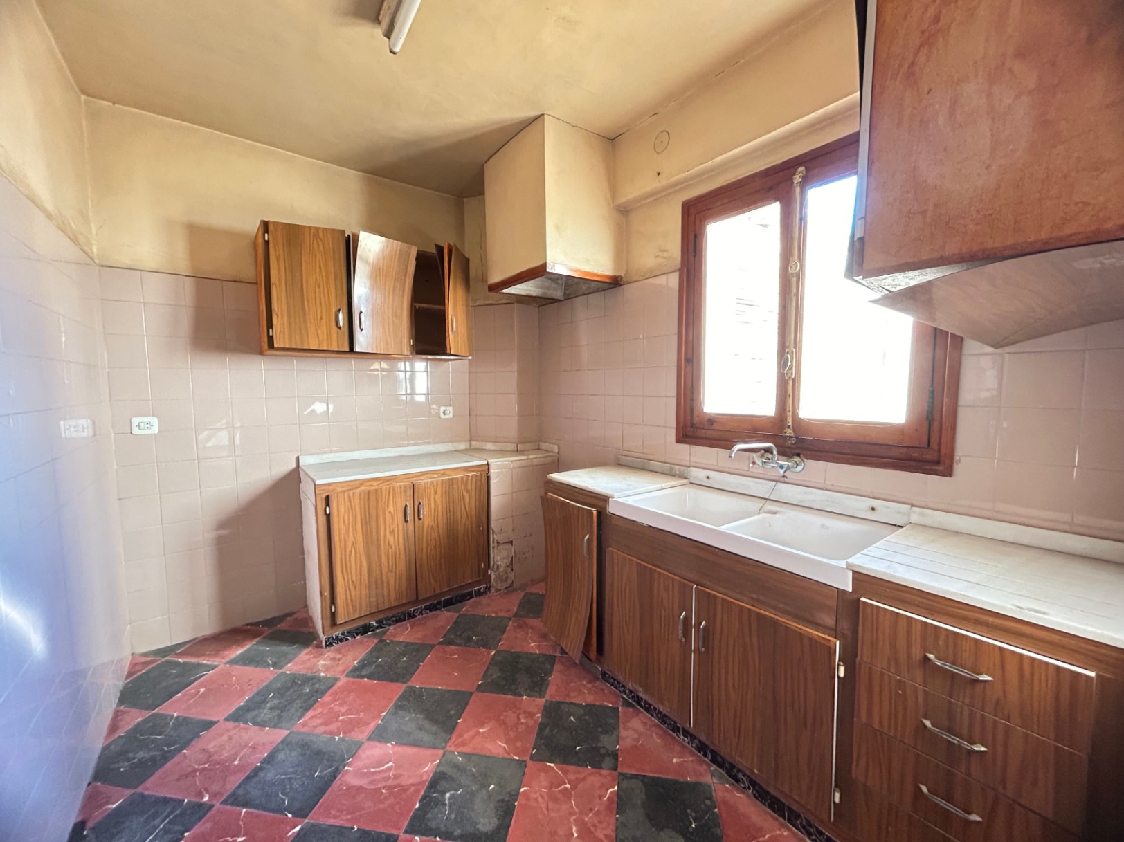 Cuisine d'origine avec sol en damier rouge et noir, meubles en bois et plan de travail en marbre blanc. Équipée d'un évier à double bac et d'une fenêtre en bois.