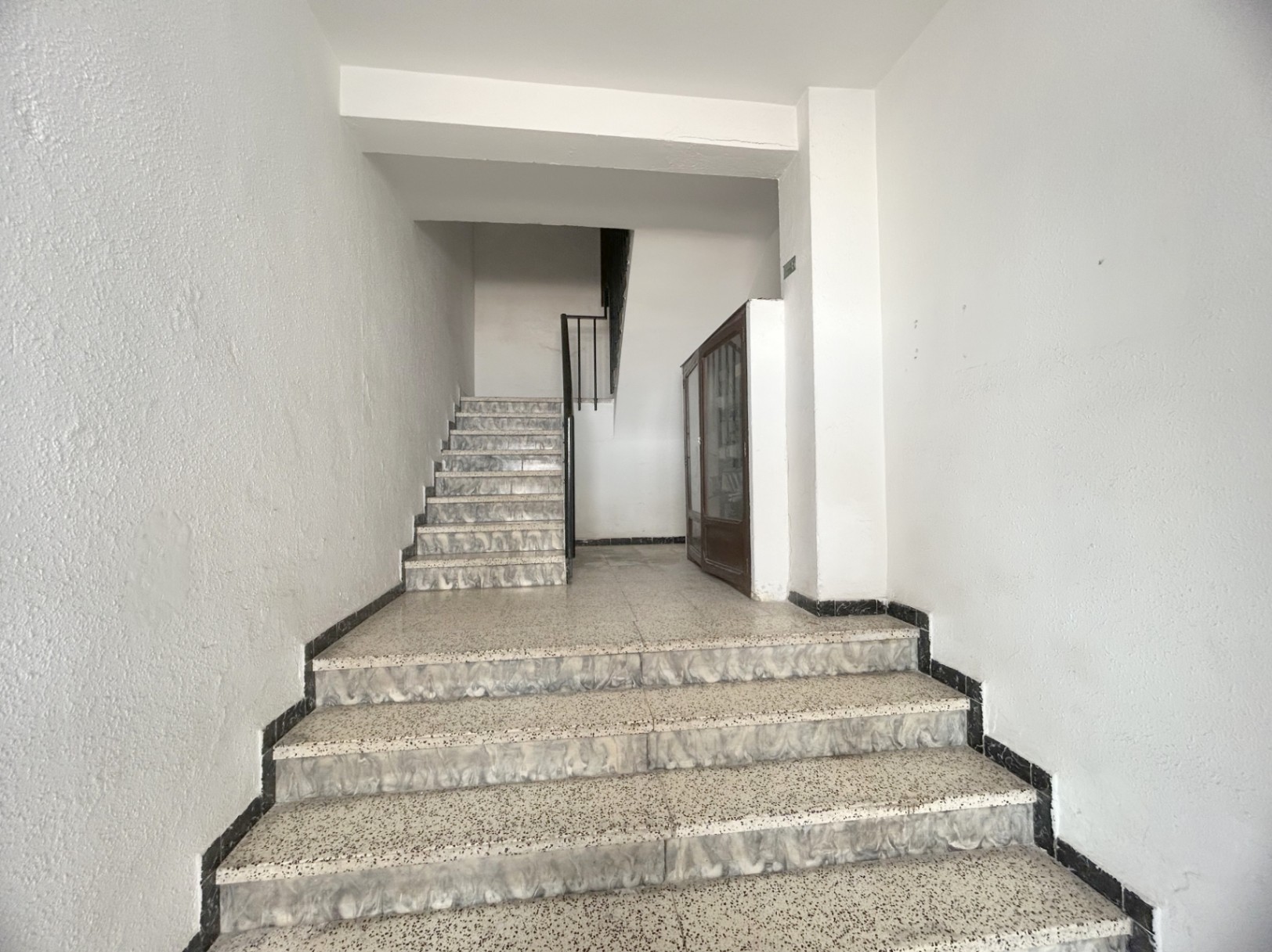 Escalier commun avec marches en terrazzo et bordures marbrées, murs blancs et rampe en métal noir. Un meuble en bois est disposé sur le palier intermédiaire.