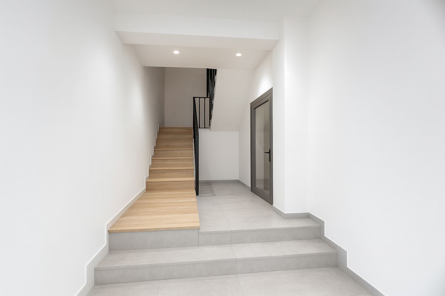 Hall d'entrée avec escalier en bois clair et rampe en métal noir. Carrelage gris au sol, murs blancs et porte vitrée avec cadre en aluminium gris.
