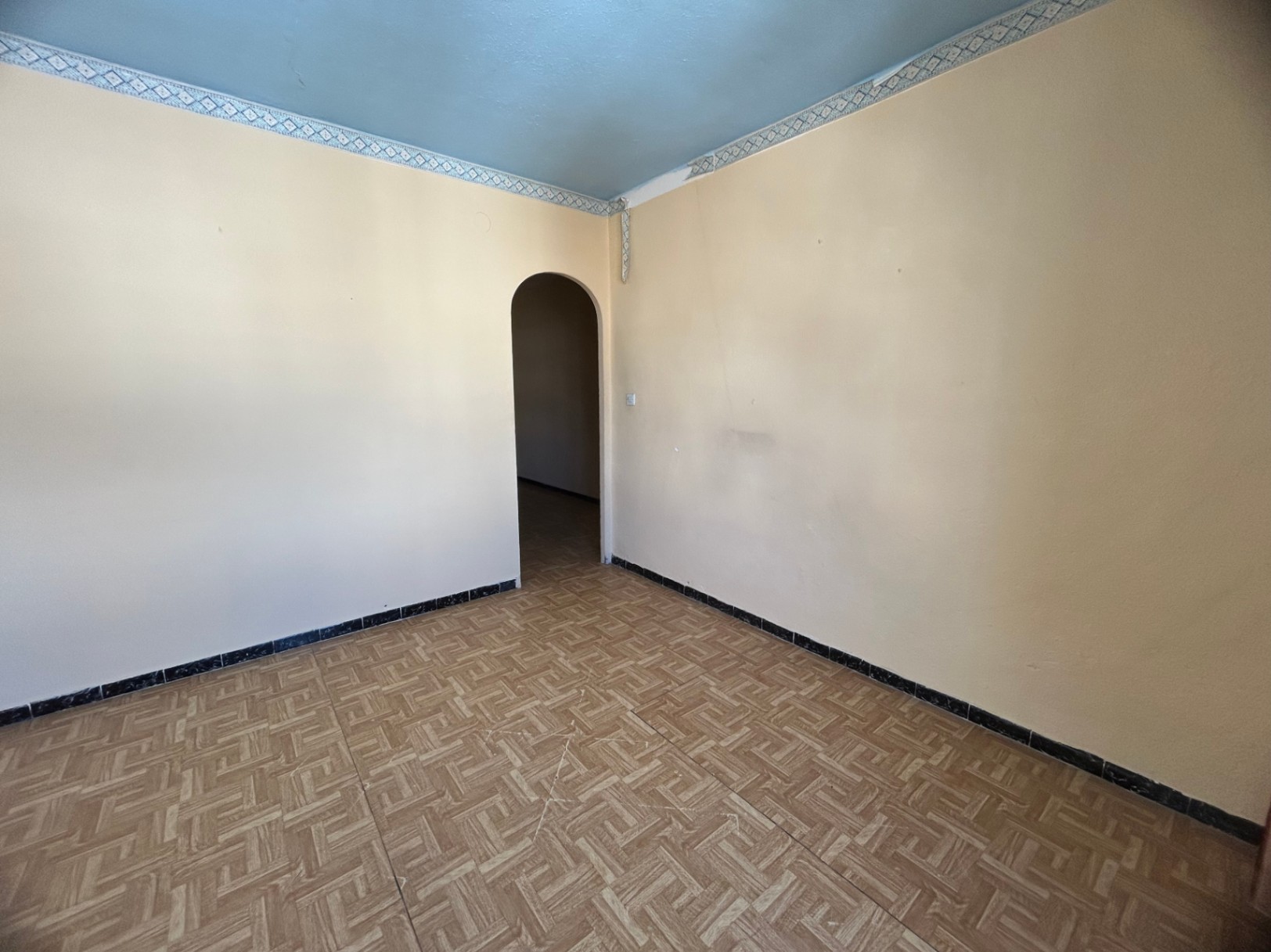 Pièce intérieure avec sol en grès cérame à motifs, murs crème et plafond bleu clair orné d'une frise. Une ouverture en arche mène à la pièce suivante.