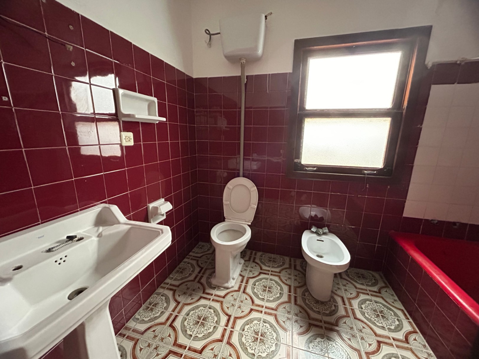 Salle de bain d'origine avec carrelage mural bordeaux, sol hydraulique à motifs et sanitaires classiques. Une fenêtre en bois assure une luminosité naturelle constante.