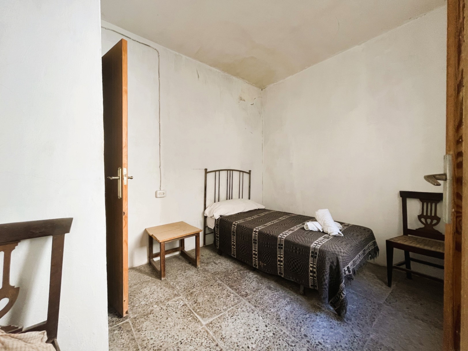 Chambre simple avec sol en terrazzo d'origine, lit individuel à tête de lit en métal et mobilier en bois traditionnel dans une pièce aux murs blancs.