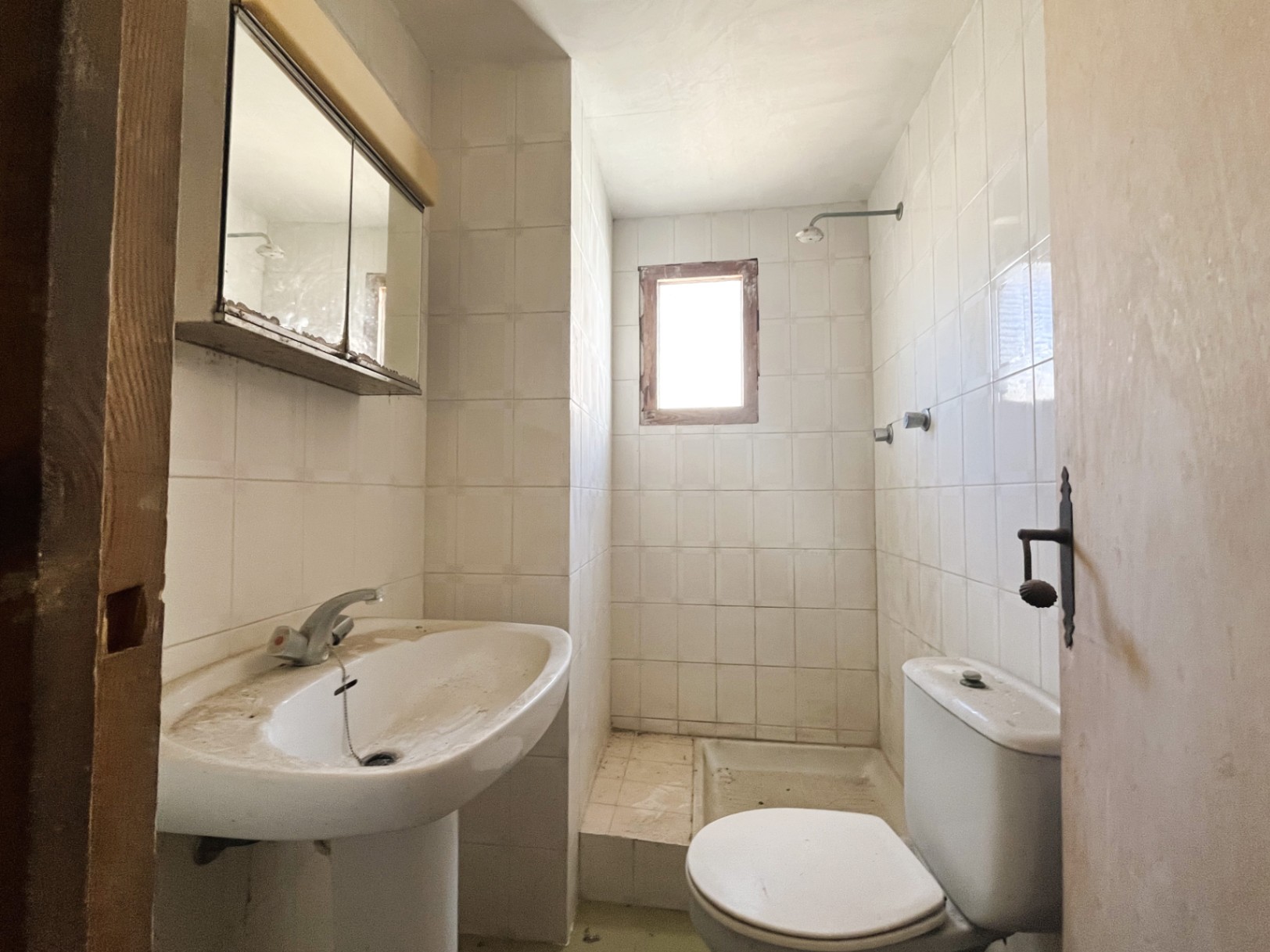 Salle de bain fonctionnelle avec carrelage blanc, lavabo sur colonne et douche avec petite fenêtre. Un espace offrant un fort potentiel de rénovation.