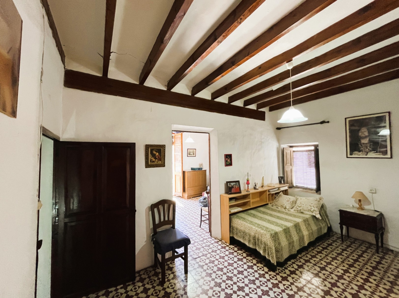 Chambre avec poutres apparentes en bois sombre, sol en carreaux de ciment d'origine et murs blancs. Elle comprend une petite fenêtre avec volets et des rangements intégrés.