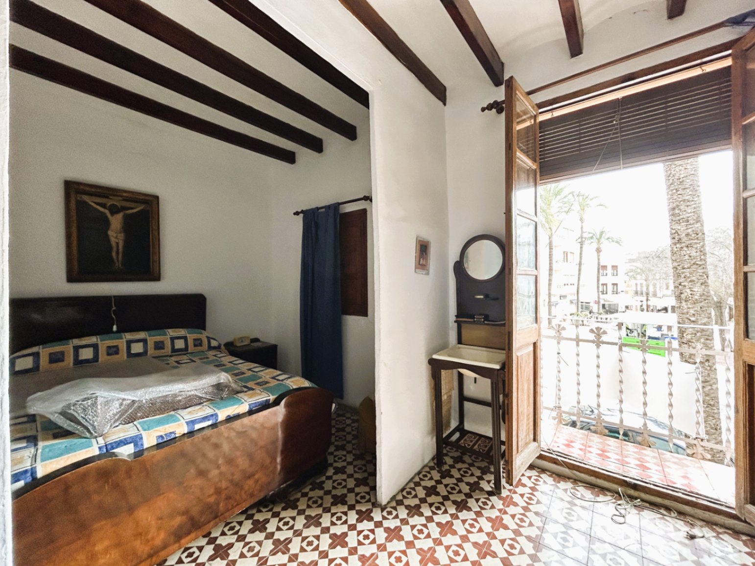 Chambre traditionnelle avec poutres apparentes, sol en carreaux de ciment d'époque et balcon offrant une vue sur la place du village et ses palmiers.
