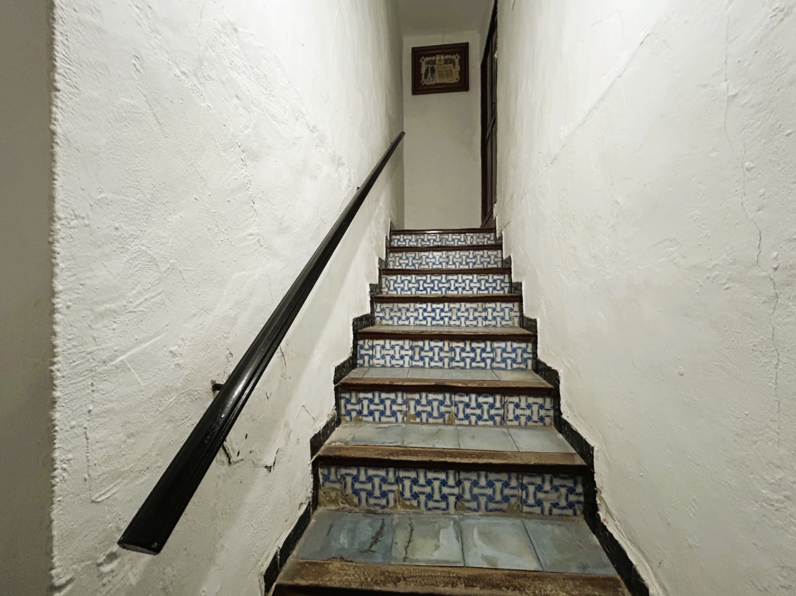 Escalier traditionnel avec contremarches en carreaux de faïence bleus et blancs, marches en bois et main courante sombre sur des murs en plâtre blanc.