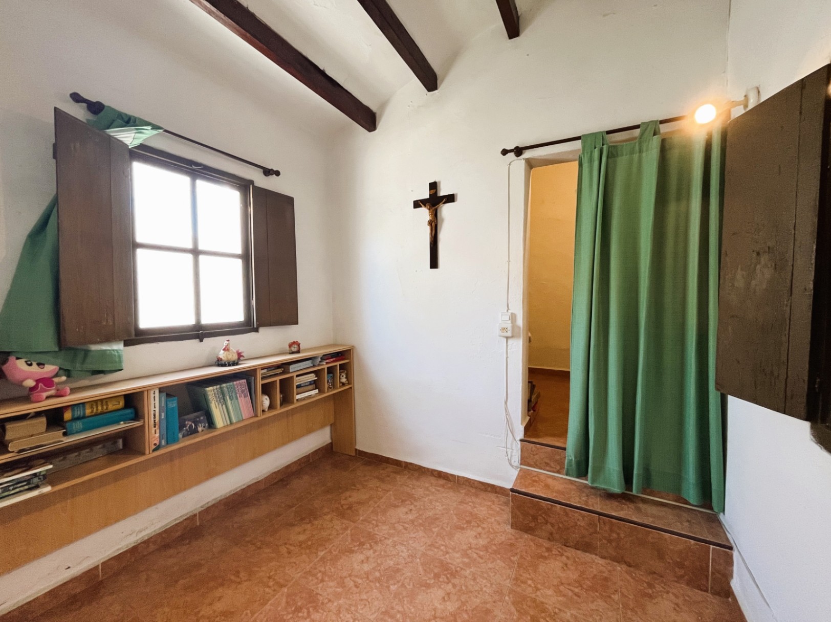 Espace intérieur avec poutres apparentes, sol en terre cuite et étagère sous fenêtre. Un accès surélevé avec marches et rideau sépare les différents niveaux de la pièce.