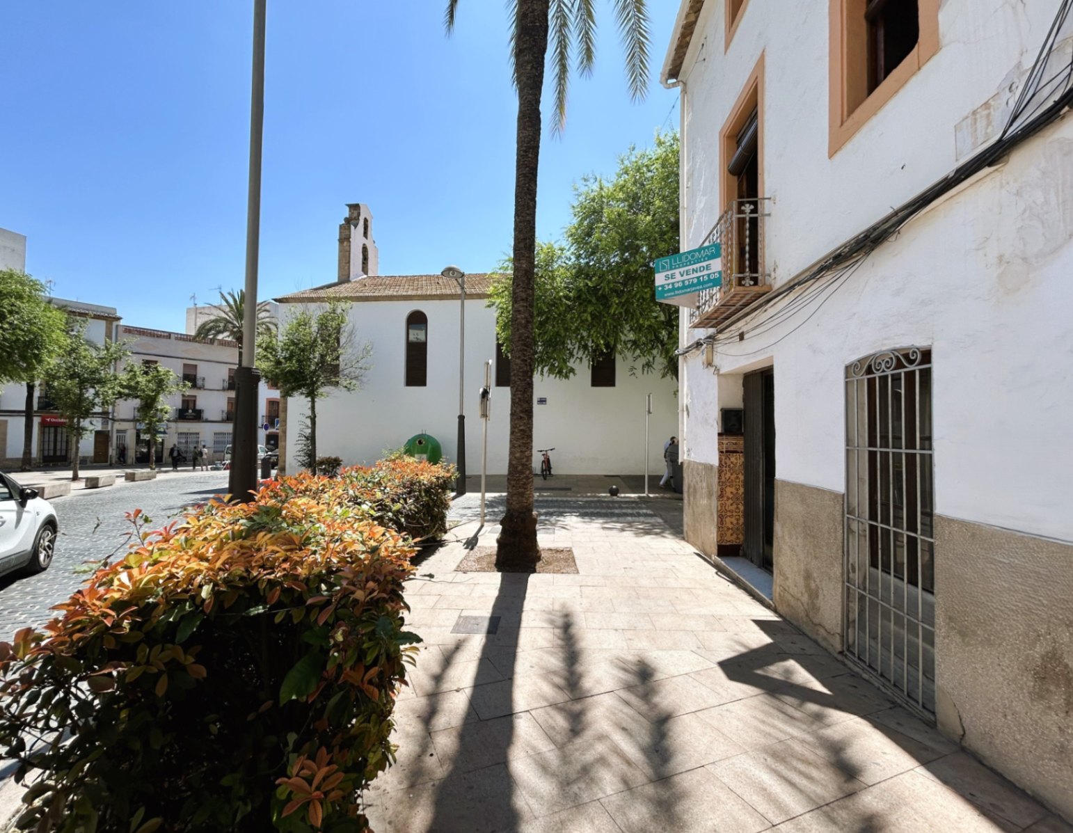 Façade blanche avec soubassement en pierre dans la vieille ville de Jávea, située à côté d'une place piétonne avec palmiers et vue sur un clocher historique.