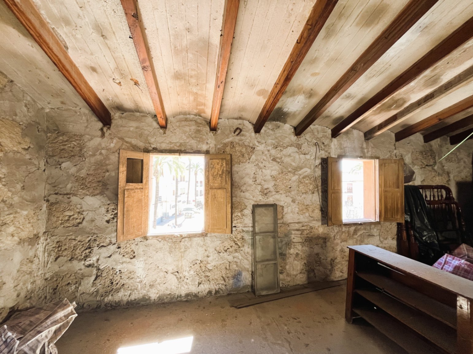 Pièce avec murs en pierre naturelle apparente et poutres en bois d'origine. Deux fenêtres avec volets en bois offrent une lumière naturelle et une vue sur la vieille ville.