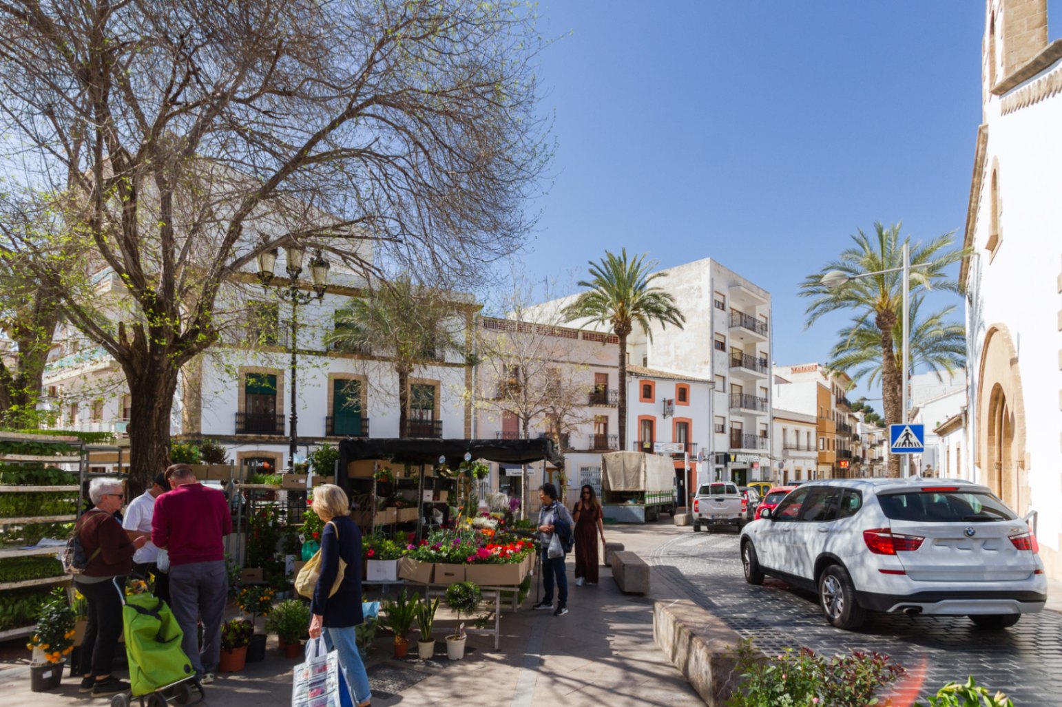 Place dans le centre historique de Jávea avec un étal de fleurs, entourée d'une architecture traditionnelle blanche, de rues pavées et de palmiers sous le soleil.
