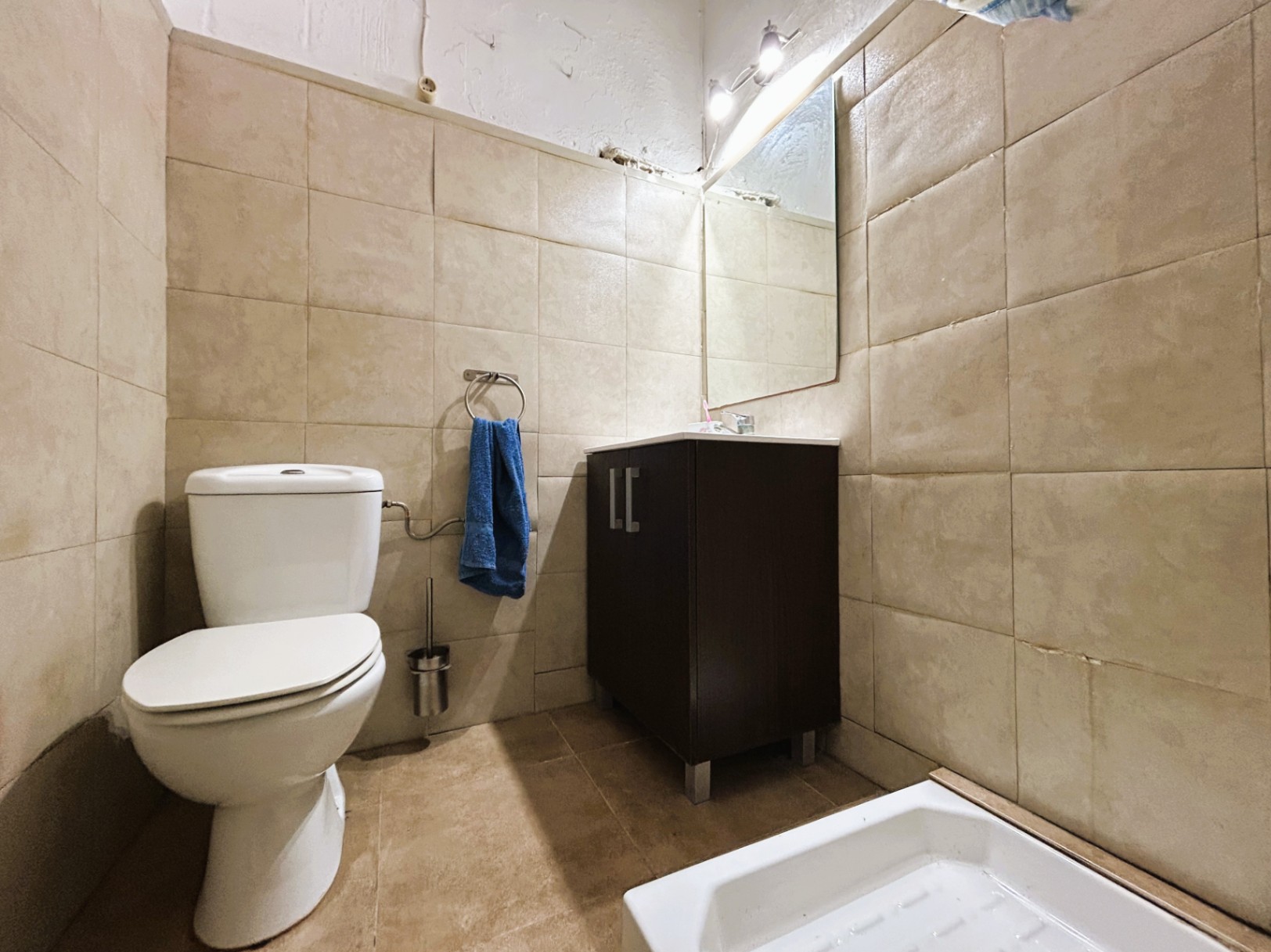 Salle de bain fonctionnelle avec murs carrelés beige, toilettes en céramique blanche et meuble vasque en bois sombre avec miroir éclairé.