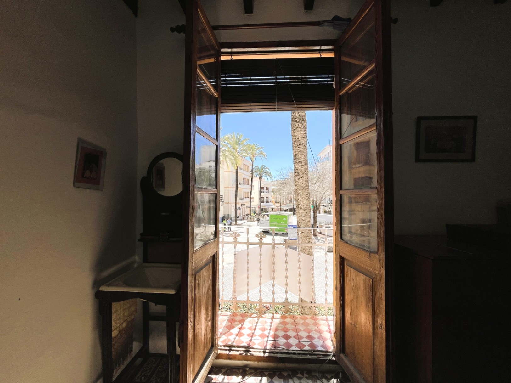 Vue intérieure à travers des portes en bois s'ouvrant sur un balcon aux carreaux rouges et blancs, surplombant une place arborée du centre historique.