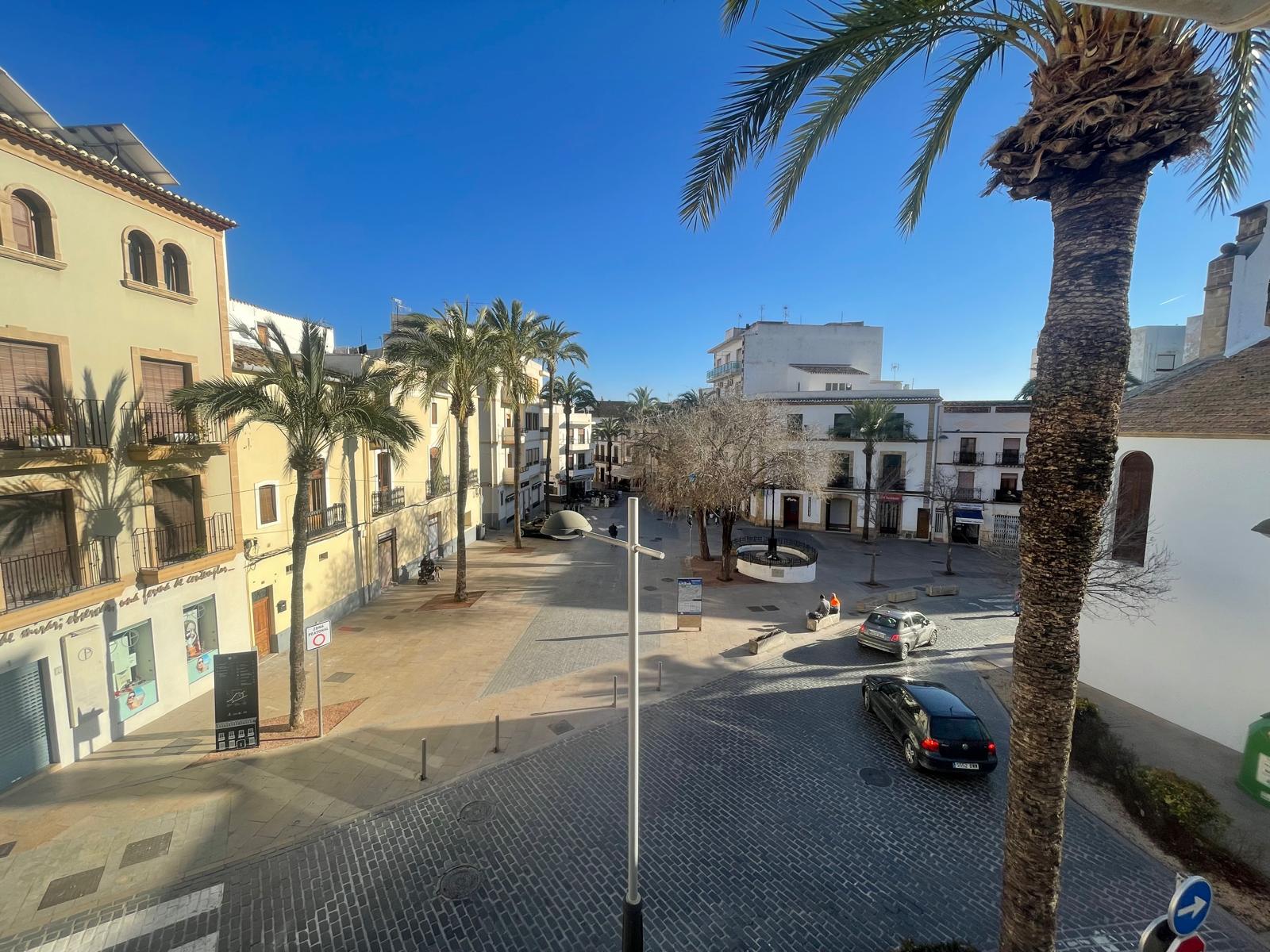Vue surélevée depuis la propriété sur une place pavée du centre historique de Jávea, bordée de bâtiments traditionnels et de palmiers.
