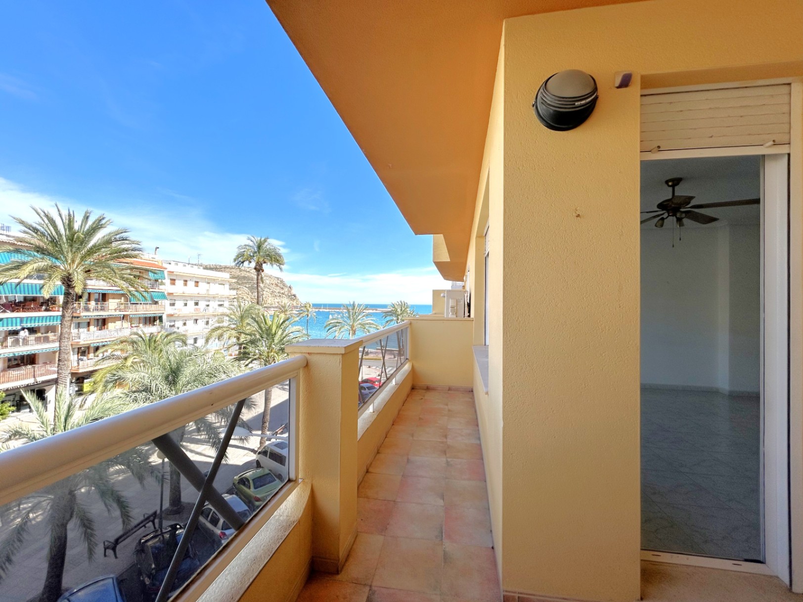 Balcon filant avec revêtement en céramique offrant une vue latérale sur la mer et le port de Jávea, entouré de palmiers et d'immeubles.